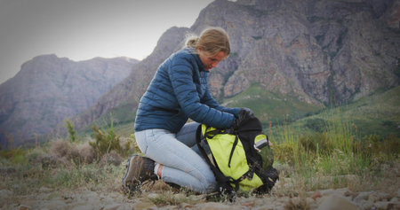 Kneeling hiker wearing navy jacket unpacking gear from neon yellow backpack in mountain valley. Adventure, outdoor, rugged, exploration, resilience, wilderness, natureの写真素材