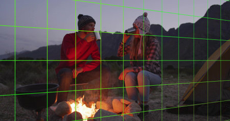 Holding metal mugs beanie-clad couple warming by ringed campfire at campsite, with cast-iron pot. Adventure, outdoor, cozy, rustic, wilderness, leisure, twilightの写真素材