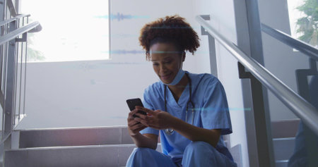 Sitting mid adult nurse in blue scrubs checking smartphone at hospital stairwell, with stethoscope. Healthcare, medical, professionalism, wellness, technology, hospital, staffの写真素材