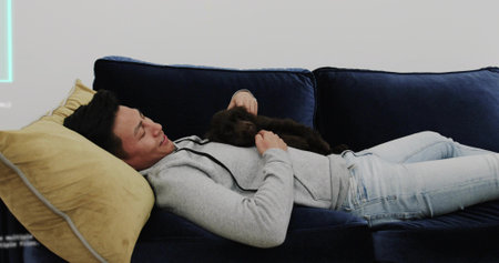 Reclining Asian male wearing gray hoodie, jeans stroking puppy on living room sofa, with neon light. Luxury, relaxation, comfort, tranquility, modern, cozy, lifestyleの写真素材