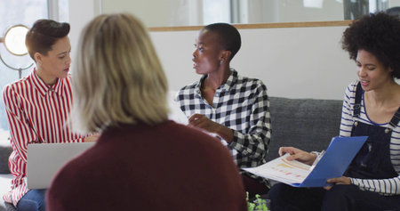 Collaborating four women sharing work materials in modern office lounge, with laptop and binder. Teamwork, collaboration, co-working, modernoffice, professional, discussion, productivityの写真素材
