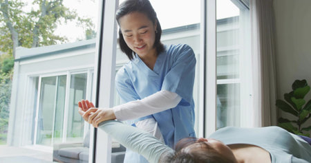Assisting therapist in blue scrubs moving patient arm on treatment table in therapy room by windows. Rehabilitation, healthcare, manual therapy, wellness, modern, relaxation, healingの写真素材