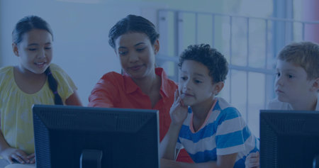 Female instructor guiding three schoolchildren in computer lab classroom, with desktop computers....
