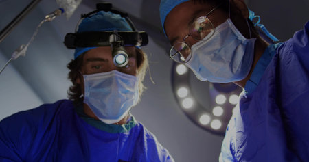 Performing operation two male surgeons focusing in operating room, with overhead surgical light....