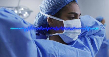 Surgeon adjusting mask straps in operating room, with blue scrubs, cap, lamp and ECG waveform....