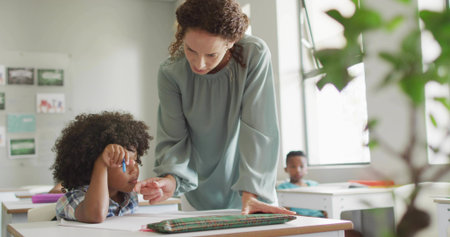 Leaning woman teacher assisting boy writing in notebook at white desk in classroom, with sunlight....