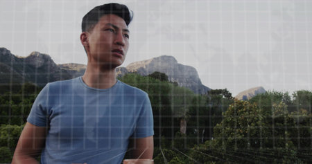 Gazing adult Asian male standing at forested mountain viewpoint, with rugged peaks rising behind. Adventure, exploration, nature, wilderness, scenic, outdoor campaign, tranquilityの写真素材