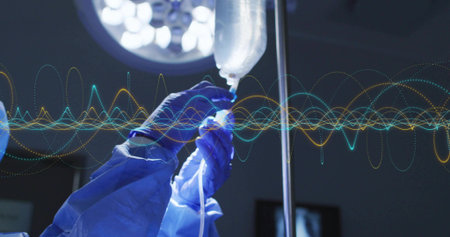 Gloved hands adjusting IV bag in operating room under surgical lamp, with roller clamp drip chamber....