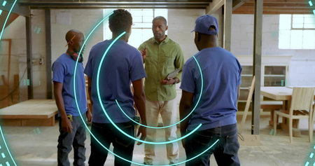Gesturing team leader showing woodworking plan in spacious shop, with tablet and furniture parts....