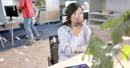 Talking Korean woman using smartphone while viewing laptop in workspace, with currency symbols. Technology, collaboration, financial, modern, professional, digital, communicationの写真素材