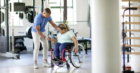 Guiding trainer steadying woman wearing active gear lifting blue dumbbell at gym, red wheelchair....