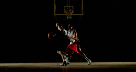 Two basketball players wearing jerseys jumping under hoop at gym free-throw area, under spotlight. Athletics, competition, adrenaline, determination, vigor, sportsmanship, dynamicの写真素材