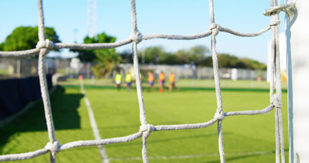 Framing white knotted goal net and right goalpost at club soccer pitch, players wearing bright bibs. Netting, goalframe, field, pinnies, outdoor, grass, teamworkの写真素材