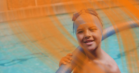 Smiling boy wearing swim cap, goggles leaning on handrail at pool with orange mesh, copy space....