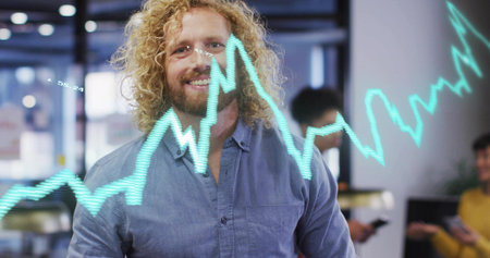 Smiling man with curly blond hair standing in modern office displaying cyan graph with smartphone. Analytics, teamwork, innovation, productivity, digital, business, collaborationの写真素材