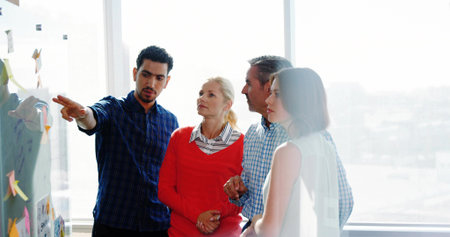 Pointing male in checkered shirt leading brainstorming at office, with sticky notes on glass board. Collaboration, innovation, teamwork, brainstorming, creativity, productivity, modernの写真素材