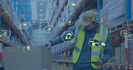 Worker scanning box with tablet in warehouse aisle, wearing safety vest and cap, with eco icons....