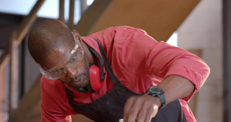 Leaning mature craftsman examining wooden plank in carpentry workshop, with goggles and earmuffs....