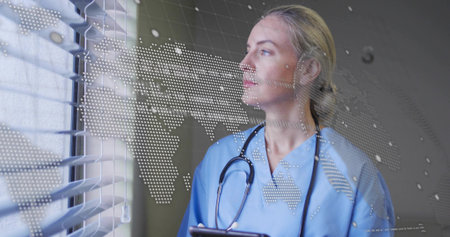 Gazing nurse holding tablet at clinic window blinds with stethoscope and dotted world map overlay. Medical, healthcare, technology, clinical, professional, innovation, analysisの写真素材