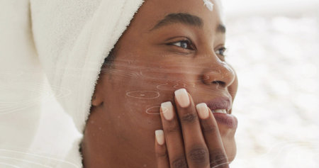 Pressing fingertips on cheek, woman applying moisturizer cream at bathroom sink, with head towel. Skincare, beauty, wellness, self-care, minimalism, elegance, tranquilityの写真素材