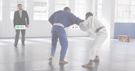 Grappling judo opponents in blue and white gis on gray mats at dojo, with referee observing. Martial...
