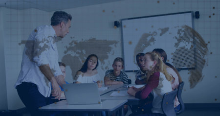 Leading male educator guiding students writing notes at classroom table, with laptop, map overlay. Mentor, collaboration, innovation, training, knowledge, digital, educationの写真素材