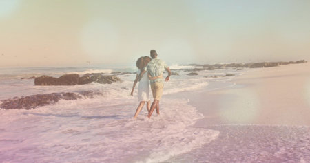 Strolling African American couple arm-in-arm into surf at sandy beach, with waves and dark rocks. Romance, relaxation, scenic, seaside, harmony, tranquility, summerの写真素材