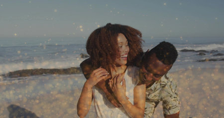 Hugging laughing couple wearing sleeveless top and tropical shirt at sandy beach, sparkling waves....