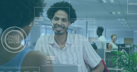 Smiling man leaning on desk talking at open-plan office with laptop, digital overlay graphics....