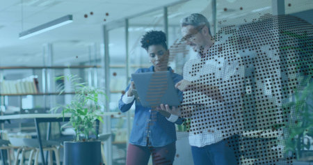 Reviewing laptop two coworkers standing behind glass partition in office, with bookshelves, plants. Collaboration, teamwork, modern, contemporary, minimalist, professional, workspaceの写真素材