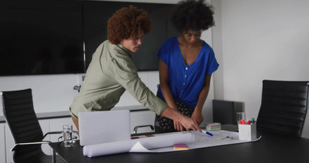 Leaning over rolled-out blueprint two colleagues collaborating at table, with laptop, sticky notes. Collaboration, teamwork, architecture, workspace, design, professionalism, modernの写真素材