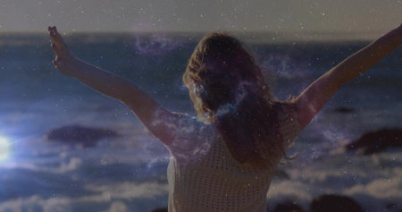 Standing woman wearing white crocheted top stretching arms at rocky shoreline sunset, ethereal mood. Serenity, freedom, tranquility, nature, bohemian, contemplative, scenicの写真素材