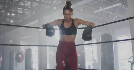 Leaning fit woman resting on boxing ring ropes at gym, showing black gloves near hanging bags....
