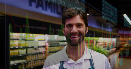 Smiling male grocery clerk wearing apron and showcasing stock charts with tickers in store aisle. Retail, commerce, analytics, data, visualization, business, technologyの写真素材