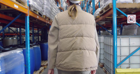 Walking warehouse worker wearing white hard hat, tan vest between tall steel racks, with blue drums....