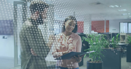 Coworkers wearing olive pink attire reviewing plan in office behind dotted glass panel, with tablet....