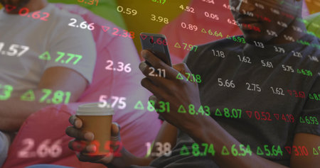 Holding paper coffee cup and smartphone, two friends watching stock market figures in living room. Casual, companionship, relaxation, modern, digital, lifestyle, comfortの写真素材