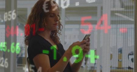 Scanning ticker on glass wall, red-haired analyst in black top using phone in office, copy space. Finance, technology, professional, digital, data, analytics, businessの写真素材