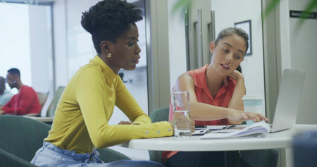 Reviewing documents, coworker women wearing mustard and coral working on laptop in office lounge....