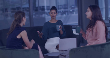 Discussing three women sharing ideas in modern office lounge, with open laptop and ceramic mugs. Collaboration, diversity, professional, conversation, workspace, modern, relaxedの写真素材