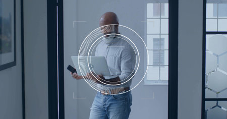 Holding laptop and smartphone, man wearing grey shirt and jeans in office, with glass partition. Professional, urban, contemporary, technology, workspace, corporate, productiveの写真素材