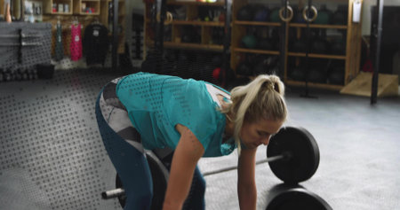 Woman wearing sportswear lifting loaded barbell in gym, with medicine balls and kettlebells....