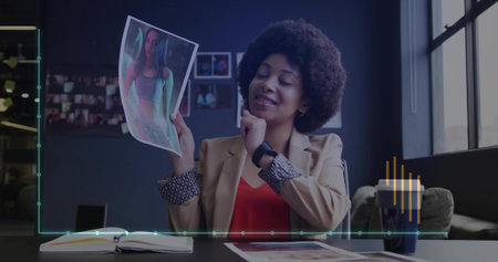 Reviewing printed picture while touching chin, woman in blazer sitting at office desk with notebook. Professional, workspace, contemporary, productivity, focus, corporate, ambianceの写真素材