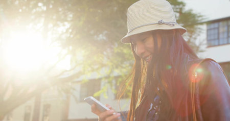 Smiling woman holding smartphone on sunlit sidewalk, striped bucket hat, leather jacket, copy space. Fashion, street style, portrait, outdoor, vibrant, casual, lifestyleの写真素材