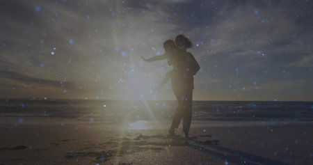 Giving child piggyback ride on sandy beach at sunset, silhouetted parent showing lens flare overlay. Family, devotion, serenity, nature, leisure, warmth, harmonyの写真素材