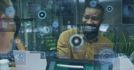 Smiling man wearing mustard top leading tech brainstorm in meeting room, laptops digital overlays. Collaboration, innovation, digital, modern, professional, teamwork, interfaceの写真素材