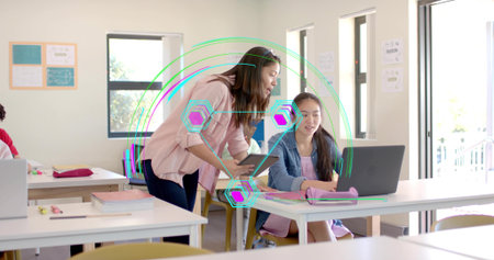 Explaining instructor leaning over student working on laptop in classroom, with tablet guidance....