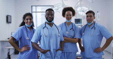 Standing group of four doctors in scrubs posing inside operating room, with stethoscopes and lights....