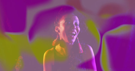 Woman with red tassel earrings and top laughing in studio, with light projections, guitar headstock....