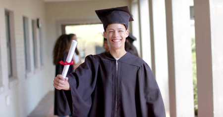 Walking students wearing black gowns and mortarboard caps down campus corridor, holding diplomas. Graduation, achievement, celebration, academic, cap and gown, success, diplomaの写真素材